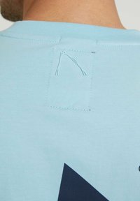 Jasnoniebieski bawełniany T-shirt z kwadratową naszywką z tyłu, odsłoniętym szwami i dużym ciemnoniebieskim nadrukiem z przodu.