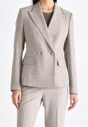 Nainen, jolla on päällään vaaleanharmaa kaksirivinen bleiseri lovikauluksilla ja siihen sopivat räätälöidyt housut, seisoo yksivärisen valkoisen taustan edessä.