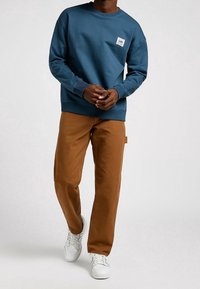 Sudadera de algodón azul con cuello redondo, que presenta un pequeño parche con un logo. Combinada con pantalones marrones y zapatillas blancas.