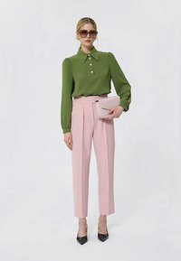 Donna con blusa verde a maniche lunghe, pantaloni rosa a vita alta, tacchi neri, che tiene una pochette rosa e occhiali da sole oversize, in piedi di fronte a uno sfondo bianco.