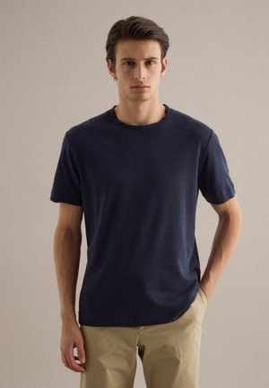 Giovane uomo con capelli scuri che indossa una maglietta blu navy a maniche corte con scollo rotondo e pantaloni beige, in piedi contro uno sfondo chiaro e semplice.