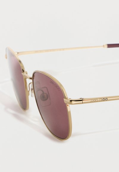 Γυαλιά ηλίου με μεταλλικό χρυσό σκελετό και φακούς με μωβ απόχρωση, με λογότυπο "Jimmy Choo" στον βραχίονα και στον φακό.