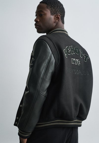 Jonge man die zijwaarts kijkt, gekleed in een zwart varsityjack met leren mouwen en "SCHOTT NYC THE KOOPLES" geborduurd op de rug.