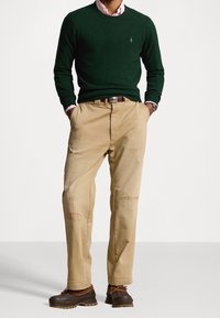 Man draagt een donkergroene trui over een roze overhemd met kraag, bruine patchworkbroeken, een bruine riem en bruine veterlaarzen, staat met handen in de zakken.