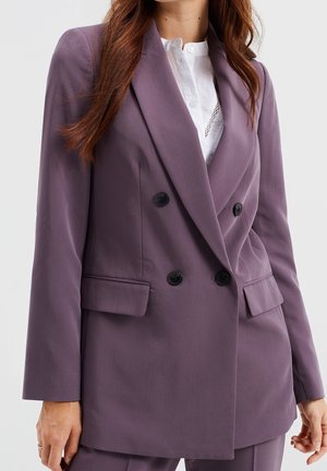 Dvojřadý blejzr z měkké, textured mauve látky. Obsahuje černé knoflíky, klopové límce a boční kapsy. Košile s detaily knoflíků pod ním.