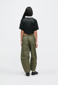 Pessoa com cabelo longo e escuro, vestindo uma camisa preta cropped, calças folgadas verdes-oliva e ténis pretos, de pé contra um fundo branco liso.