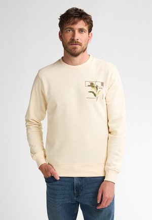 Hombre vestido con sudadera crema con gráfico de planta en el pecho y jeans azules, de pie con una mano en el bolsillo frente a un fondo liso.