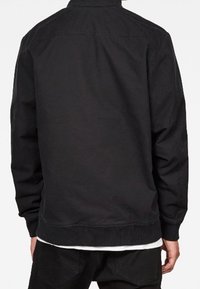 Man med svart långärmad jacka och svarta byxor, stående med ryggen mot kameran mot en enfärgad bakgrund.