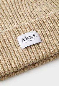 Bonnet en tricot beige avec un motif à côtes texturé. Présente une étiquette en tissu blanc avec "ARKK COPENHAGEN" imprimé en noir.