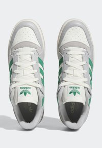 Bílé a šedé tenisky Adidas se zelenými pruhy, bílými šňůrkami a perforovanými špičkami, viděné shora na světlém pozadí.