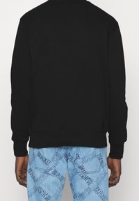 Schwarzer Pullover mit rundem Ausschnitt und langen Ärmeln, ausgestattet mit gerippten Bündchen und Saum, kombiniert mit hellblauen Jeans mit Textmuster.