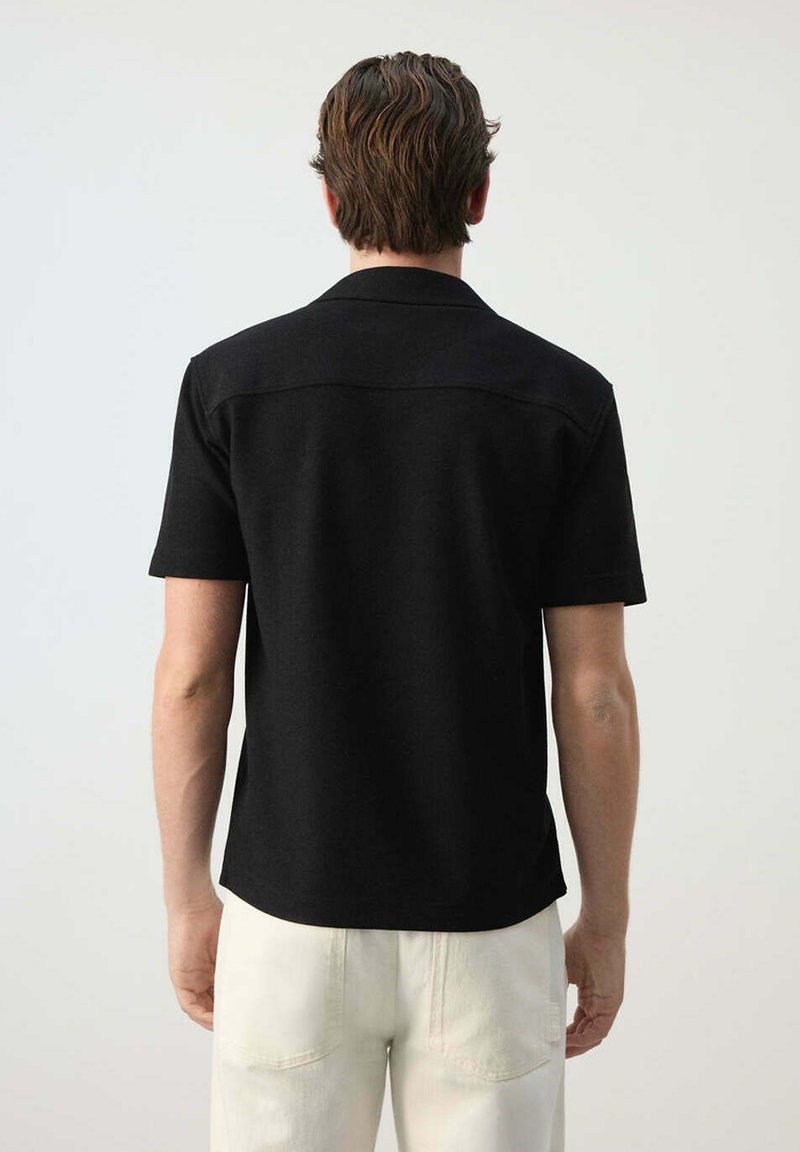 Hombre con cabello corto y castaño, vestido con una camiseta negra lisa de manga corta y pantalones blancos, de pie frente a un fondo claro y liso.