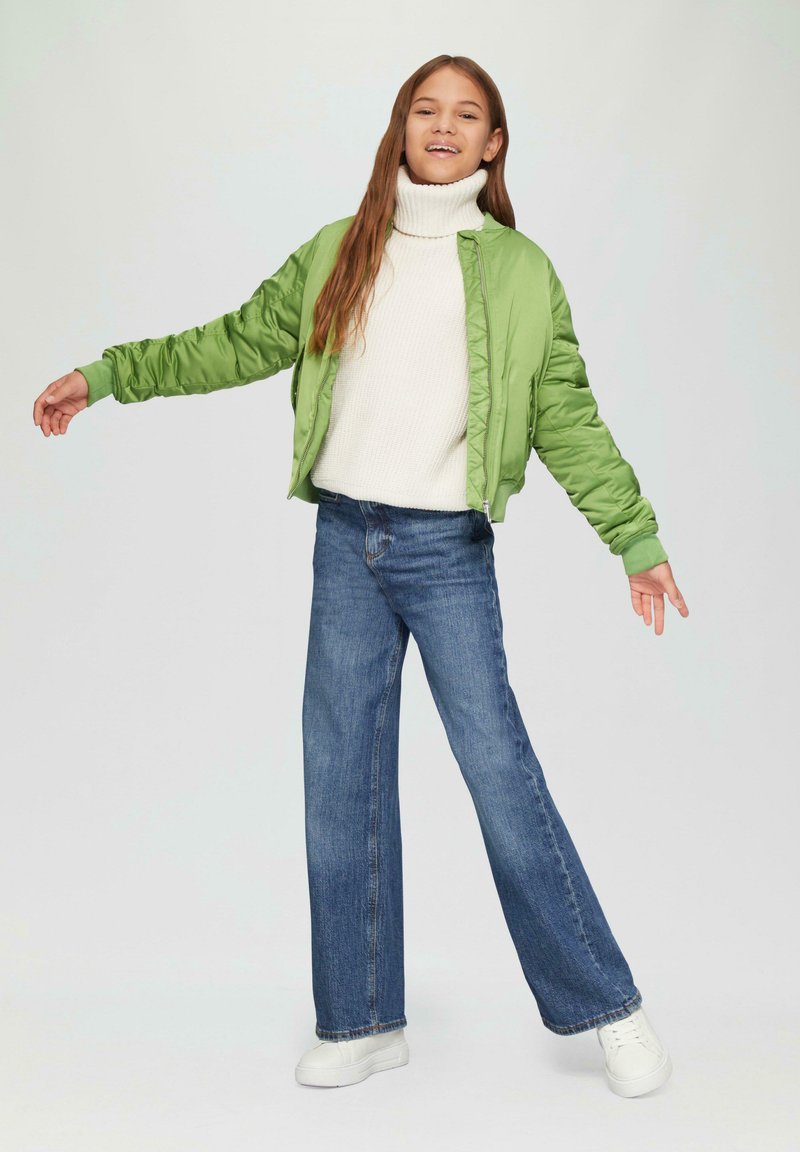 Grüne, leichte Steppjacke, weißer Rollkragenpullover und weit geschnittene blaue Jeans. Modell steht mit ausgestreckten Armen und trägt weiße Sneaker.