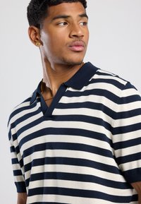 Marineblau- und cremegestreiftes kurzärmeliges Polohemd mit V-Ausschnitt und strukturiertem Strick. Akzente sind ein klassischer Kragen und eng anliegende Ärmel.