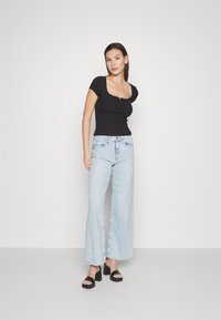 Top nero aderente con scollatura quadrata, abbinato a jeans a gamba larga blu chiaro. La modella indossa sandali neri con tacco a blocco.