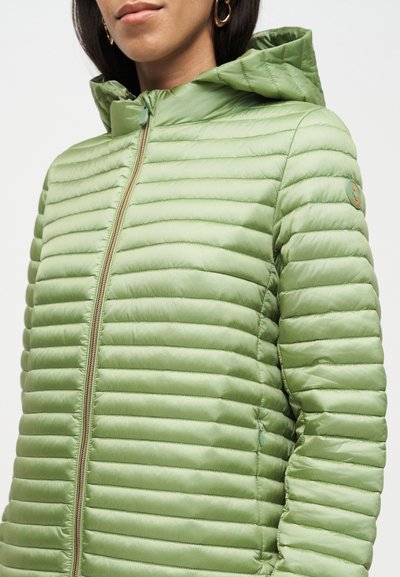 Femme portant une veste matelassée vert clair avec capuche, fermeture éclair à l'avant et un écusson rond sur la manche supérieure, sur un fond blanc uni.