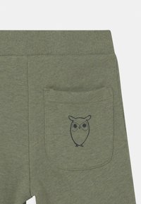 Grüne Stoffhosen mit einem gerippten Taillenbund und einer Gesäßtasche, die eine einfache schwarze Umrisszeichnung einer Eule zeigt.