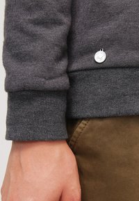 Felpa grigia con polsini a coste, trama morbida e un piccolo emblema metallico sull'orlo. Abbinata a pantaloni marroni con texture.