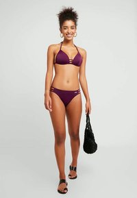 Biquíni roxo com um top triangular com detalhes em tiras e parte de baixo combinando com recortes. Modelo segurando uma bolsa tecida preta e usando sandálias.