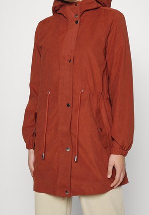 Giacca con cappuccio di colore ruggine con bottoni a pressione frontali, coulisse regolabili in vita, polsini elastici e tasche con pattina, indossata sopra pantaloni chiari.