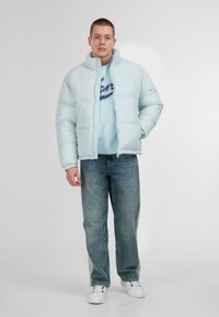 Šviesiai mėlyna pūkinė striukė su aukštu apykakle, dėvima ant šviesiai mėlyno džemperio su užrašu, derinama su laisvo kirpimo džinsais ir baltais sportbačiais.