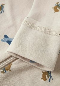 Tissu beige clair présentant une texture douce, avec un motif animal subtil en bleu et marron. Le design des poignets montre des détails de couture soignés.