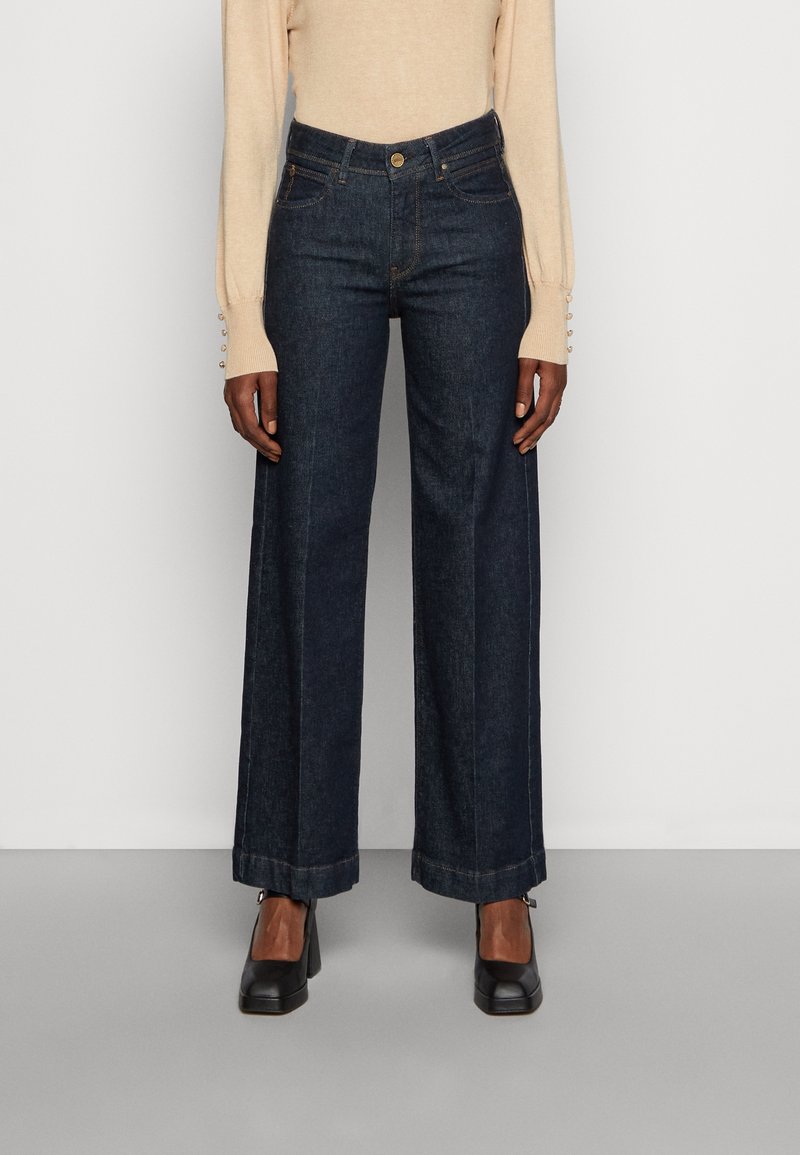 Kvinna iförd mörkblå vida jeans, svarta klackskor och en beige långärmad topp med knäppta manschetter, stående mot en vit bakgrund.