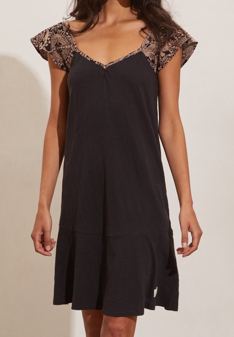 Femme portant une robe noire sans manches avec des manches courtes à motif floral beige et un décolleté en V.