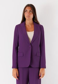 Blazer viola con rever a lancia, chiusura a un bottone e due tasche frontali. Tessuto liscio con una vestibilità sartoriale.
