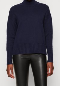 Maglione lavorato a maglia blu navy con colletto alto, polsini e orlo a coste, e una vestibilità rilassata. Indossato con pantaloni neri a vita alta.