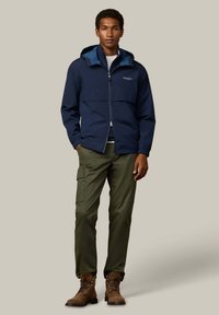 Tmavomodrá vetrovka so zapínaním na zips, elastickými rukávmi a kapucňou, v kombinácii so zelenými cargo nohavicami a hnedými šnurovacími topánkami.