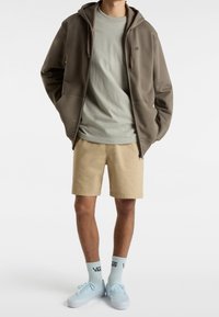 Brązowy polar z zamkiem na całej długości nałożony na jasnoszary t-shirt, w połączeniu z beżowymi szortami. Jasnoniebieskie sneakersy oraz białe skarpetki z czarnym logo dopełniają całość.