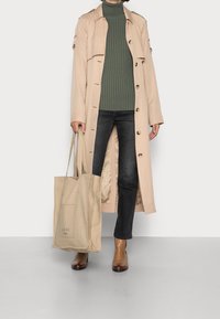 Person, die einen beigen Trenchcoat, einen grünen Rollkragenpullover, dunkle Jeans und braune Stiefeletten trägt und eine beigefarbene Tragetasche mit Aufdruck dabei hat.