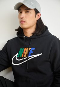Felpa nera con logo "NIKE" colorato; presenta uno swoosh bianco, cordini e una tasca frontale. Abbinata a un cappellino grigio chiaro.