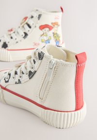 Baskets montantes blanches pour enfants avec motif vache noir, accents rouges, fermeture éclair latérale, et personnages Woody et Jessie de Toy Story imprimés à l'arrière.