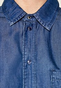 Jeansskjorta med detaljer såsom klassisk krage, framknappar i nyanser av blått och en ficka på vänster sida med synlig sömnad.