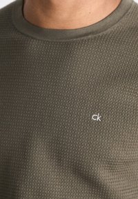 Olivgrüner Pullover mit einer strukturierten Musterung, Rundhalsausschnitt und weißem "ck"-Logo auf der linken Brustseite. Weicher Stoff mit einem lässigen Design.
