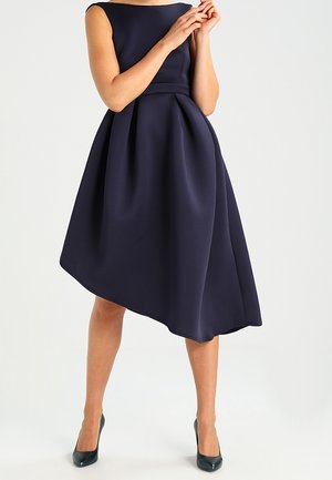 Laivastonsininen A-linjainen mekko, jossa hihaton malli, kerätty vyötärö ja epäsymmetrinen helma. Sileä kangas, jossa jäsennelty siluetti ja minimaalinen koristelu.