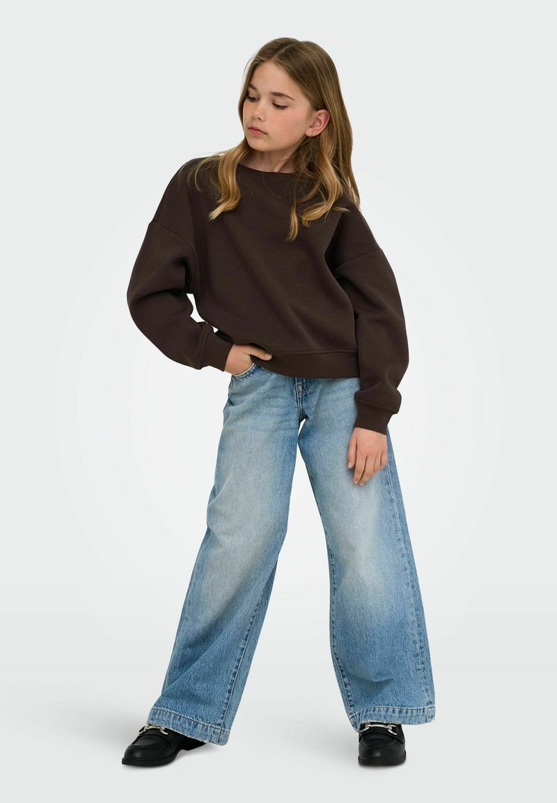 Jeune fille portant un sweat-shirt oversize marron foncé, un jean large bleu clair et des chaussures noires, debout avec une main dans la poche.