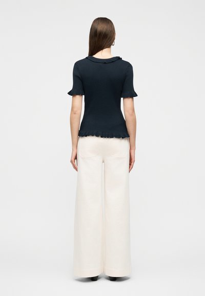 Γυναίκα με μακριά καστανά μαλλιά, φορώντας εφαρμοστή μαύρη κοντομάνικη μπλούζα με βολάν και λευκό παντελόνι με φαρδιά μπατζάκια, στέκεται με την πλάτη στραμμένη.