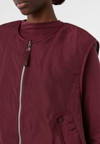 Burgundowa bezrękawnik z zamkiem z przodu, wykonany z gładkiego materiału. Posiada boczną kieszeń z guzikiem oraz warstwowy design z wysokim kołnierzem.