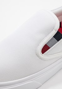 Weiße Slip-On-Sneaker aus strukturiertem Canvas, mit einem roten und marineblauen gestreiften Akzent an der Seite und einer weißen Gummisohle.
