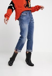 Personne portant un jean bleu usé retroussé, des bottines noires, et un pull rouge ample avec des accents de manches noir et blanc.