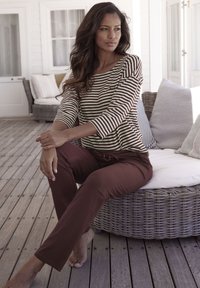 Top de manga larga a rayas en marrón y crema, combinado con pantalones burdeos. Modelo sentado en muebles de mimbre, descalzo sobre una terraza de madera.