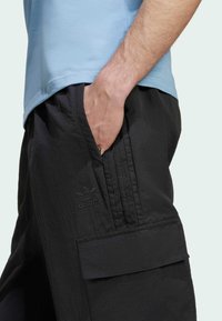 Czarne szorty cargo o teksturze z haftowanym logo Adidas. Posiadają kieszeń boczną i plisowany krój. Noszone z jasnoniebieską koszulą.