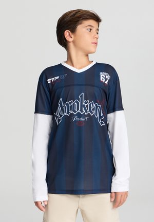 Ragazzo con indosso una maglietta sportiva a righe blu navy e nera sopra una camicia a maniche lunghe bianca, pantaloni beige, che guarda di lato su uno sfondo neutro.