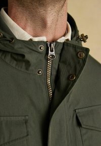 Alyvuogių žalios spalvos striukė su aukšta apykakle, metaline užtrauktuku ir užsegimais. Turi priekinės kišenes ir patvarų kanvos tekst�ūrą.