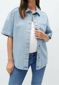 Camisa chaqueta de denim azul claro, de mangas cortas, con botón en el frente, un bolsillo en el pecho, corte relajado y un dobladillo ligeramente deshilachado.