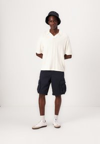 Polo à manches courtes en tricot crème, shorts cargo navy avec poches latérales, et baskets blanches avec des accents gris, portées avec un chapeau seau noir.