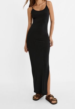 Femme portant une robe longue noire ajustée avec des bretelles fines et des sandales à boucle imprimé léopard, debout devant un fond blanc.